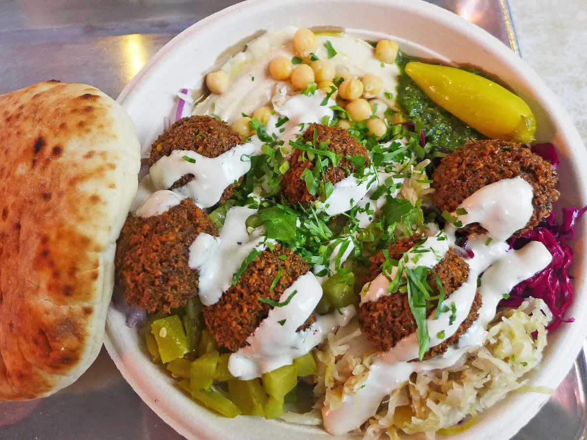 A round container with falafel, pickles, hummus, purple cabbage, etc., with puffy pita on the side.